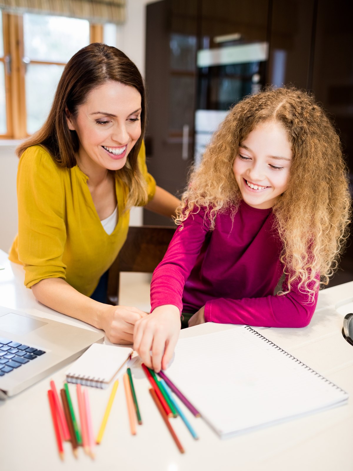 A woman leaning in beside a girl at a table spread with coloured pencils and an open notebook